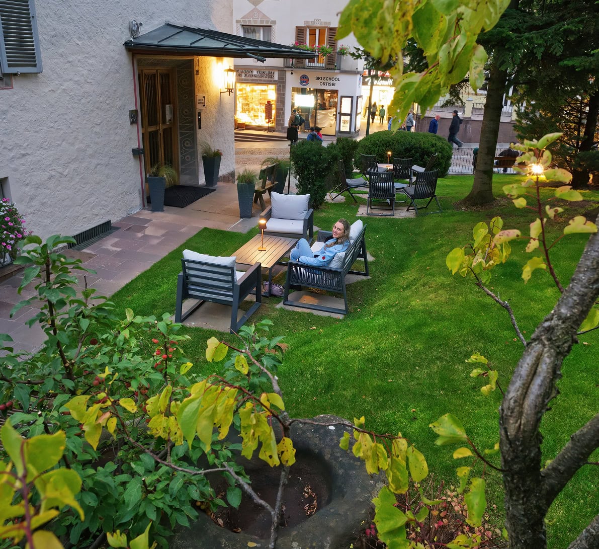 Aquila Dolomites Residence: Ortisei Below, the Dolomites Beyond, and You in Between 20 The garden area at Aquila Dolomites with a woman sitting on an outdoor sofa.