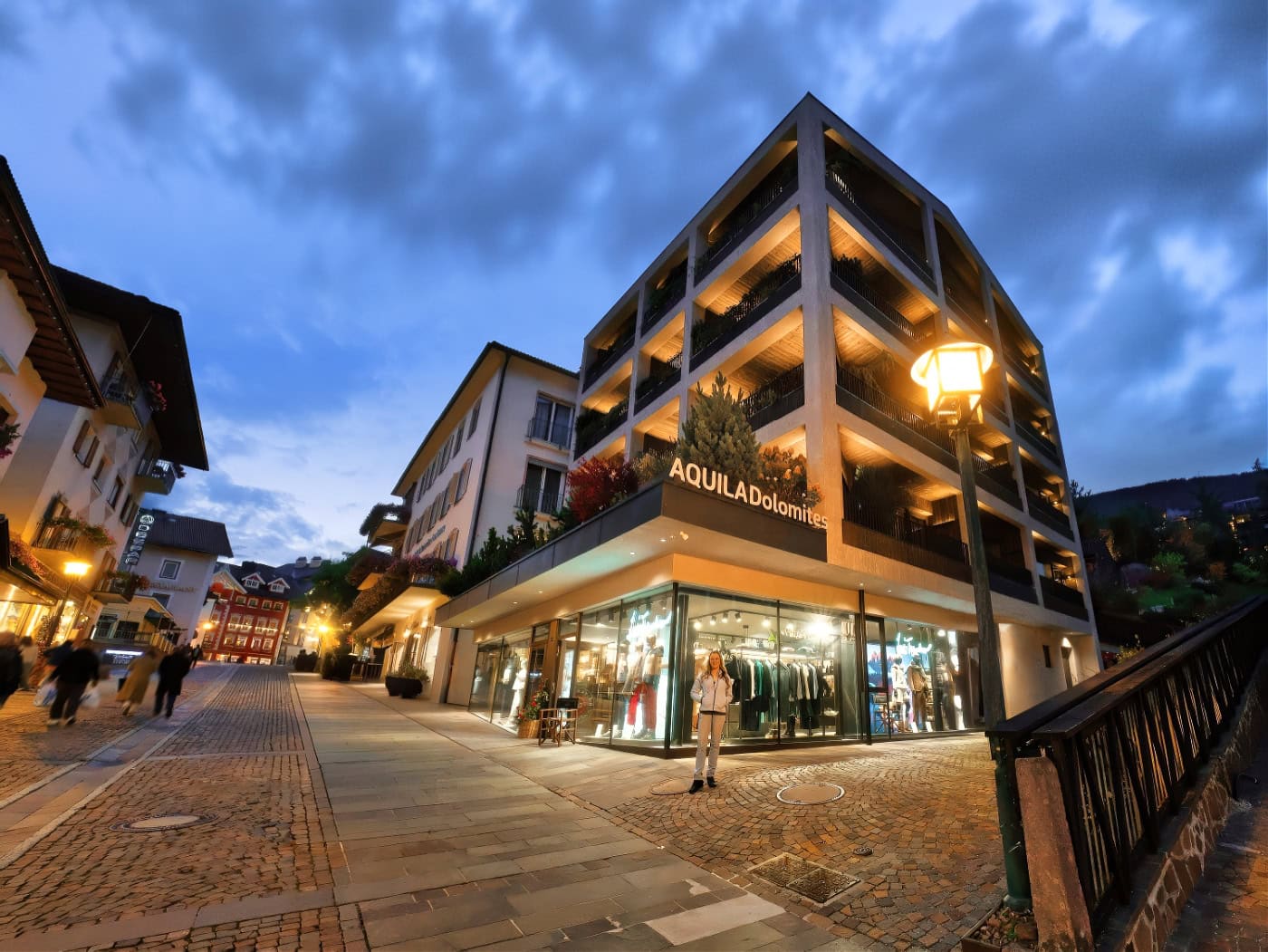 Aquila Dolomites Residence: Ortisei Below, the Dolomites Beyond, and You in Between 3 A woman standing in front of Aquila Dolomites Residence in Ortisei at dusk.