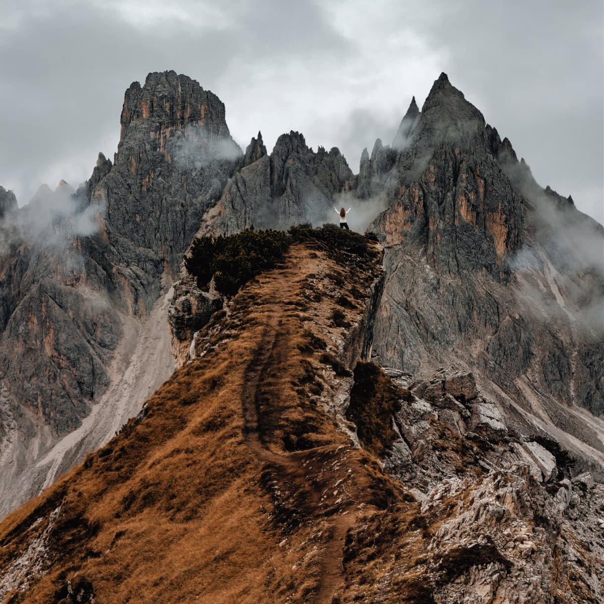 A female hiker standing with arms raised at the Cadini di Misurina viewpoint on a gloomy October day.
