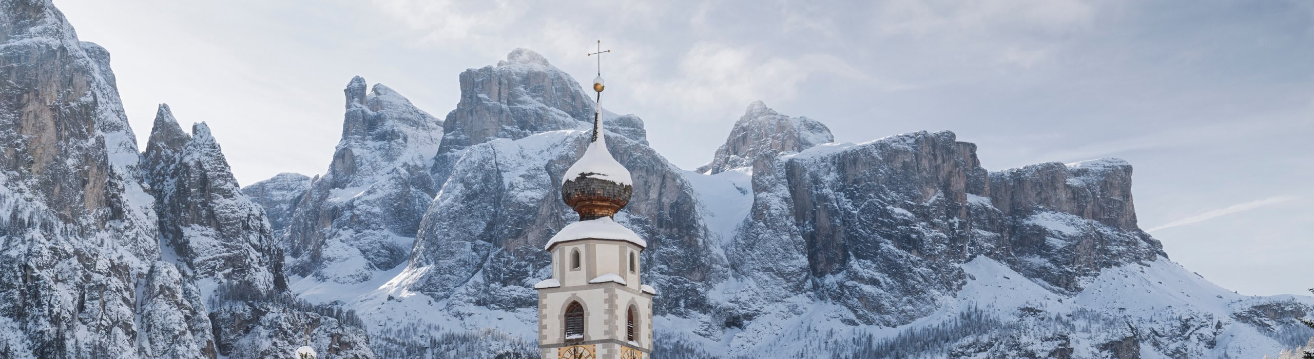The steeple of a the church in Colfosco in Alta Badia with snow-covered Dolomite mountains in the background.